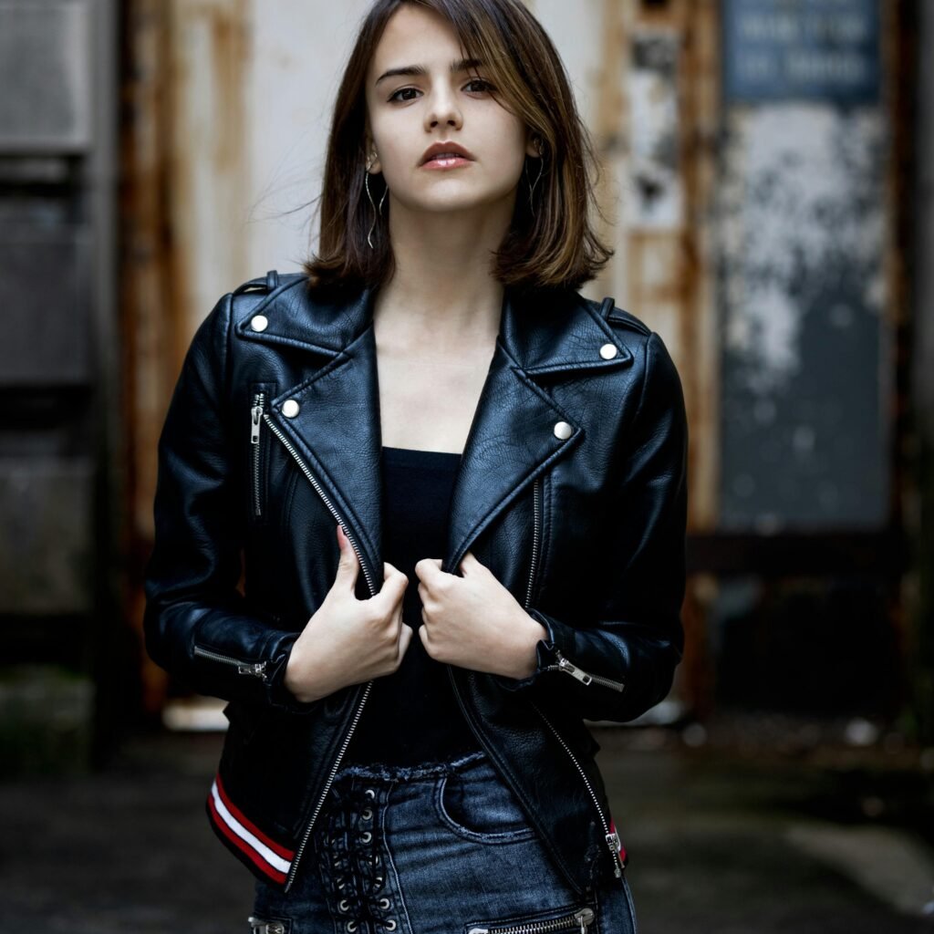 A young woman stands outdoors wearing a black leather biker jacket over a black top, looking confidently at the camera.
