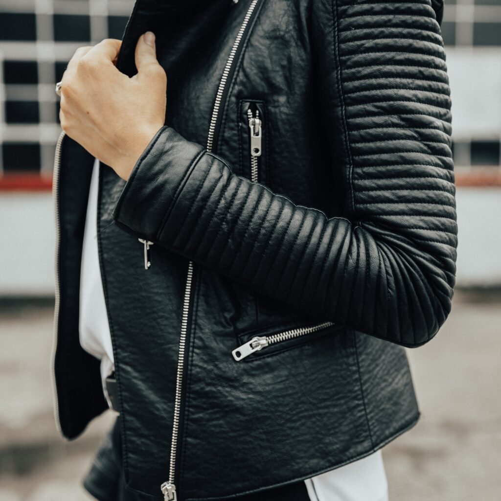 A close-up side view of a person wearing a black quilted leather biker jacket with metal zippers and a white outfit underneath.