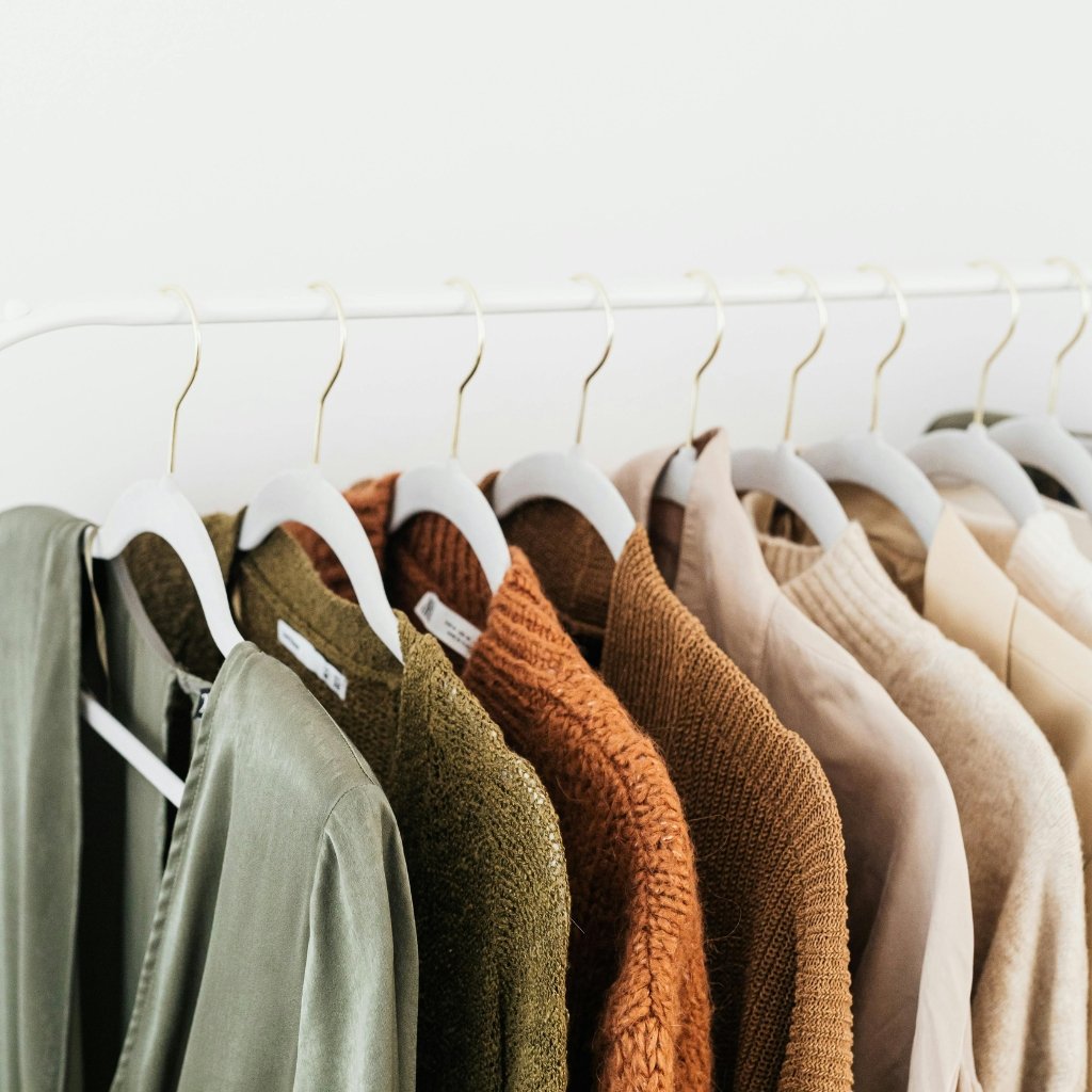 Row of coats and sweaters in earthy tones hanging on white hangers along a clothing rack.