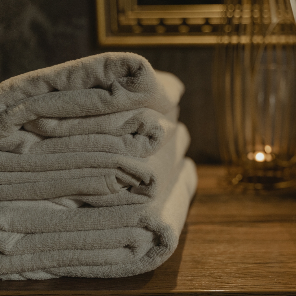 A neatly folded stack of light gray towels on a wooden surface with a warm lamp in the background.