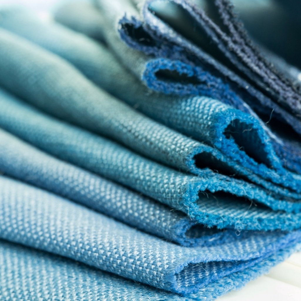 A stack of folded blue denim fabrics and jeans with varying shades of blue, showing texture and frayed edges.

