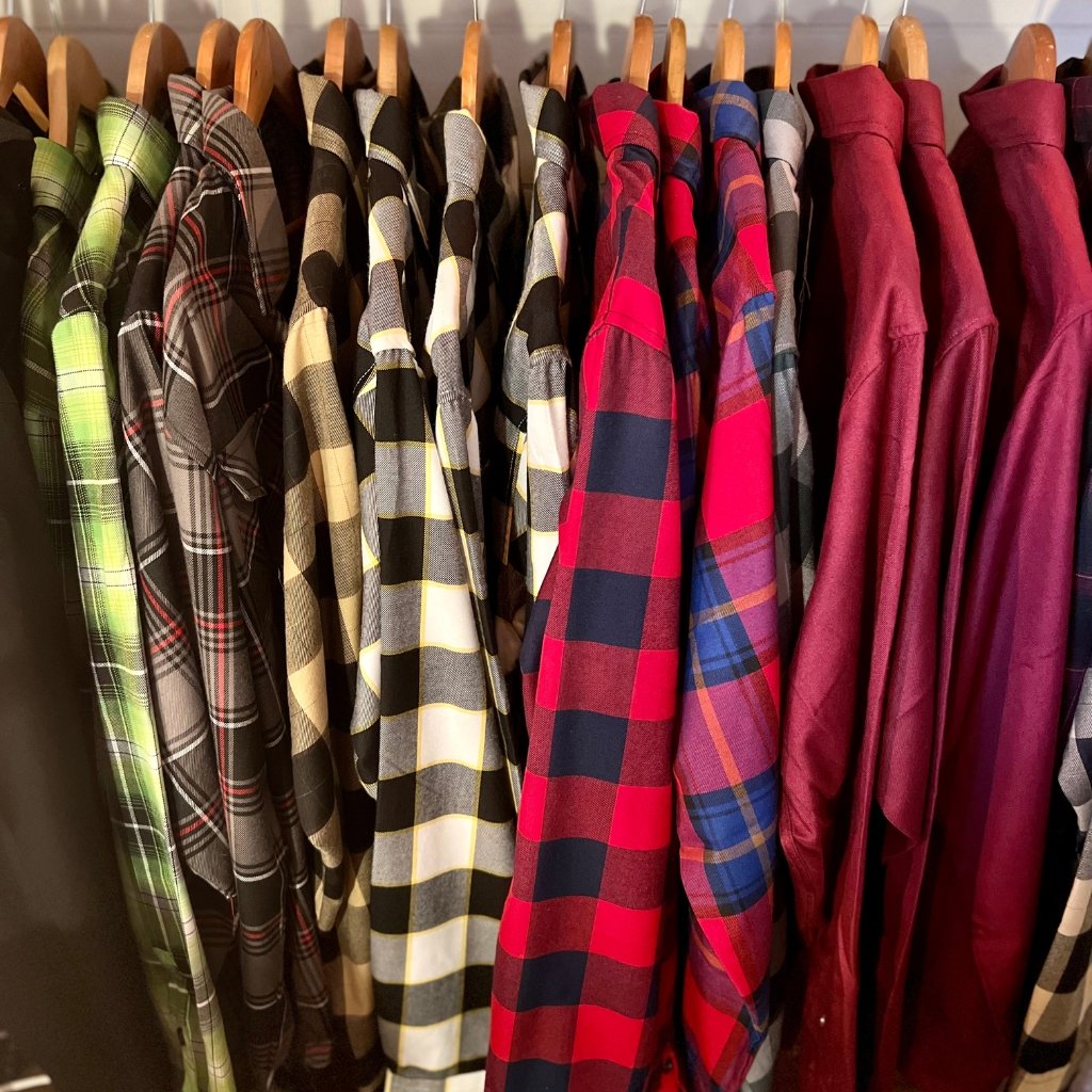  A row of plaid and solid-colored shirts on hangers in a closet, displaying a range of patterns including green, black, beige, red, and maroon.

