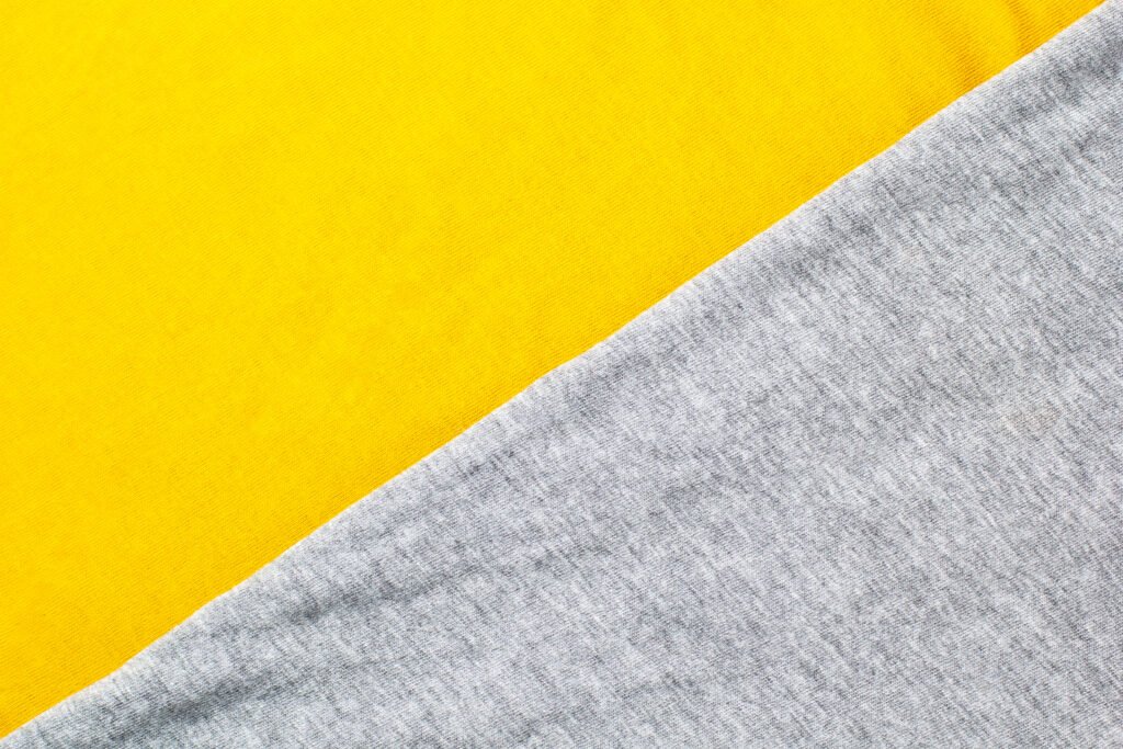 Close-up view of two folded fabric layers: a bright yellow rayon fabric on top and a heather gray cotton fabric beneath, with a diagonal seam where the two textures meet.