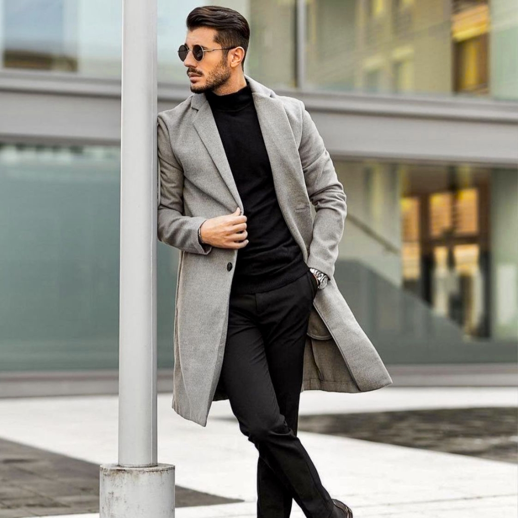 Chesterfield Overcoat Guide Classic Style with Modern Appeal 7 A stylish man stands outdoors beside a grey lamppost, wearing a long light grey overcoat, a black turtleneck, black trousers, and dark sunglasses. He leans against the post with one hand in his pocket and the other adjusting his coat, showcasing a modern, urban look.