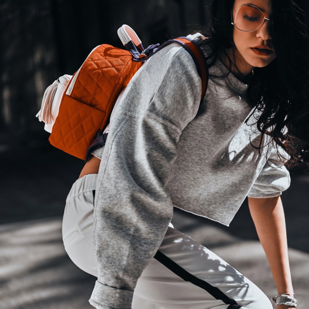 A stylish woman wearing a gray cropped sweatshirt and white pants with a black stripe, riding a bike while carrying an orange quilted backpack.