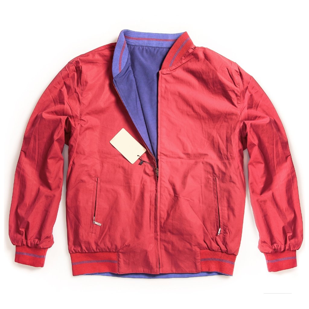 A red bomber jacket laid flat on a white background. The jacket has blue lining, ribbed cuffs and hem in red and blue, and zippered pockets. A white price tag hangs from the zipper pull.