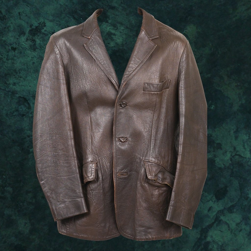 A brown leather workwear field jacket displayed against a dark green textured backdrop, featuring a notch lapel, two front pockets with flaps, a chest pocket, and three-button front closure.