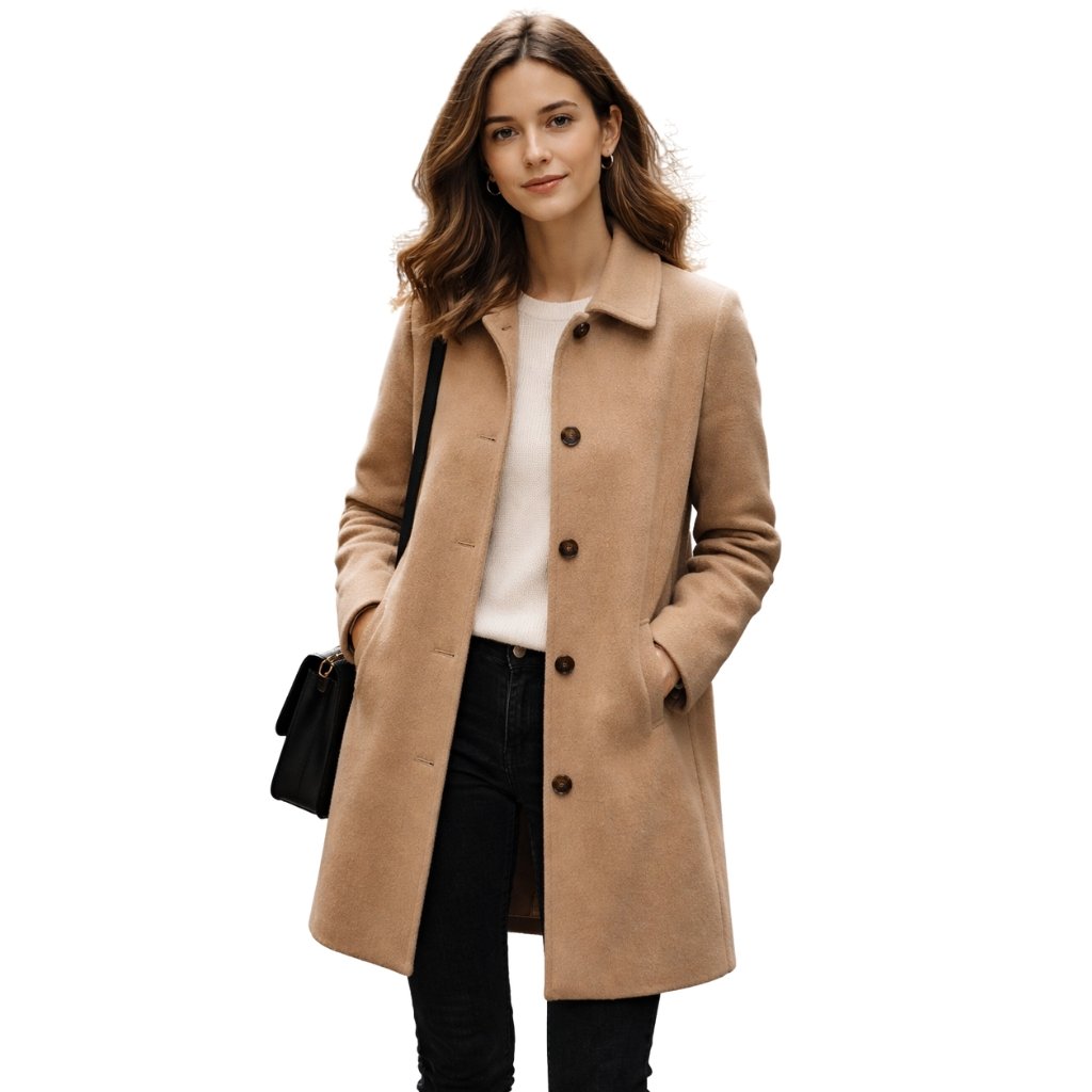 A woman stands wearing a camel-colored knee-length coat over a light sweater, with hands in pockets and a black shoulder bag.