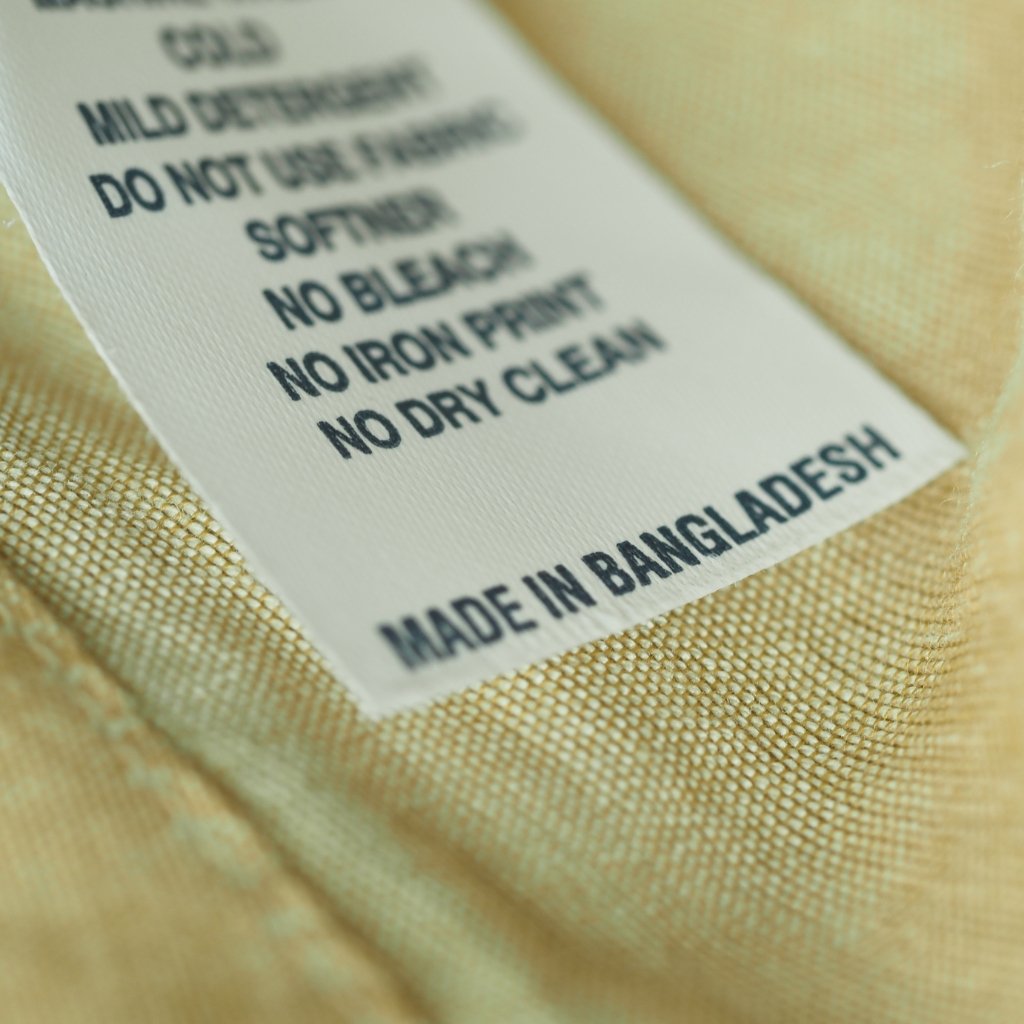 A close-up of a fabric care label sewn into a light-colored garment. The label shows printed care instructions and ends with “MADE IN BANGLADESH.”


