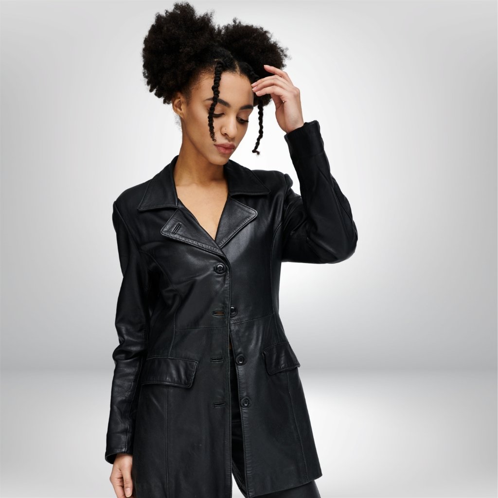 A stylish Black longline leather jacket worn by a woman with natural hair. The jacket has a wide lapel, button closure, and flap pockets, falling to mid-thigh length. The model poses with one hand in her hair against a neutral gray background.
