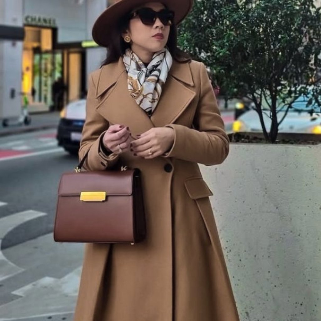A stylish woman stands on a city street wearing a camel-colored long wool coat, a printed scarf, large sunglasses, and a wide-brimmed hat. She carries a structured brown handbag with a gold clasp.