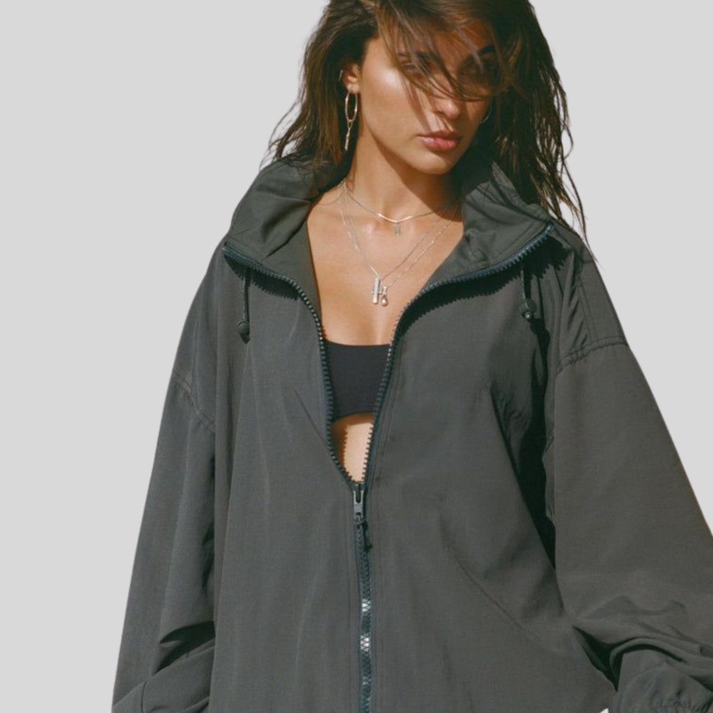 A woman wearing a dark oversized zip-up jacket with a black top underneath, against a light gray background. Her hair partially covers her face, and she is accessorized with layered necklaces and hoop earrings.