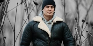 Classic Leather Parka Jacket for Men’s Stylish Outerwear A man standing outdoors wearing a dark green leather parka with a cream shearling collar, gray hoodie underneath, and a black beanie. He is in a muted, desaturated background with tall, thin plants.