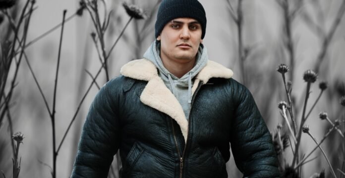 A man standing outdoors wearing a dark green leather parka with a cream shearling collar, gray hoodie underneath, and a black beanie. He is in a muted, desaturated background with tall, thin plants.