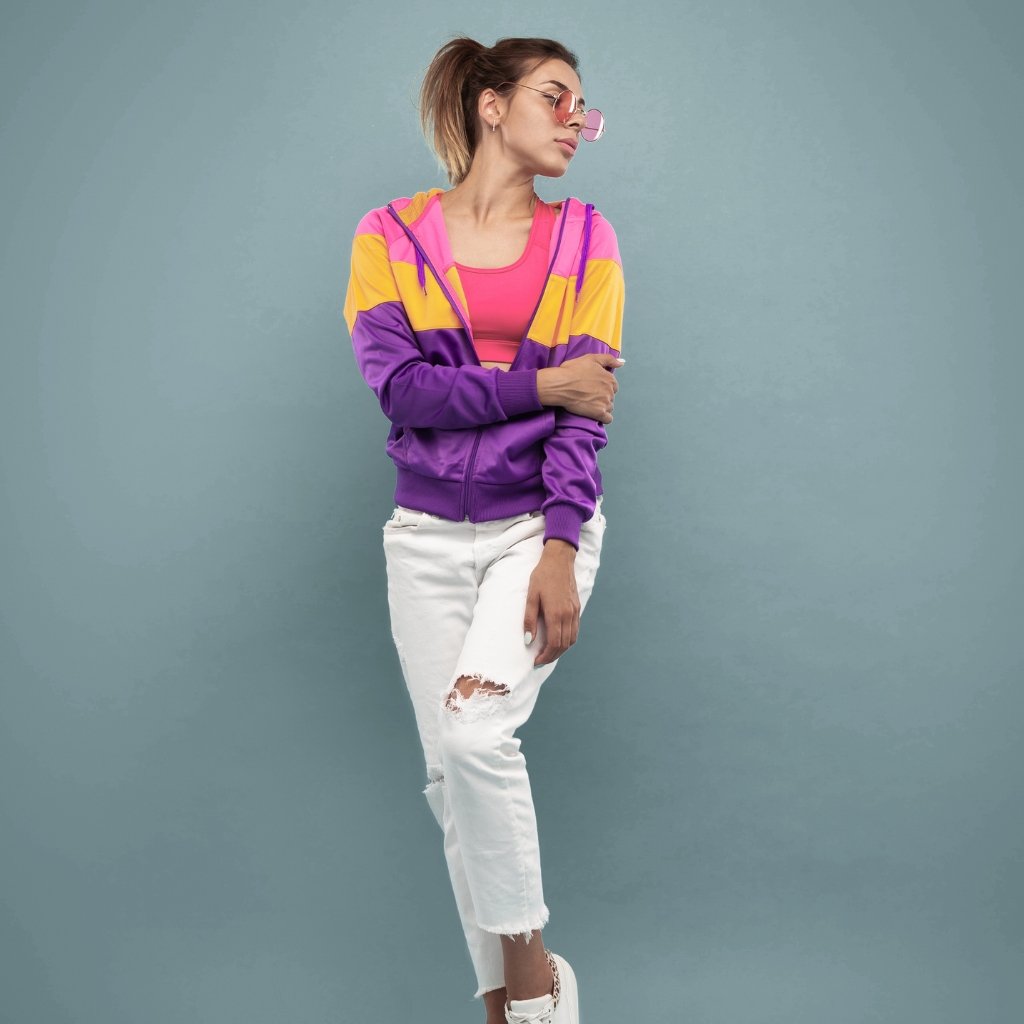 A stylish young woman stands against a teal wall, wearing a purple, pink, and yellow color-block zip-up hoodie, a pink top, white ripped jeans, and white sneakers. She has sunglasses and her hair in a ponytail, posed with one arm across her body.
