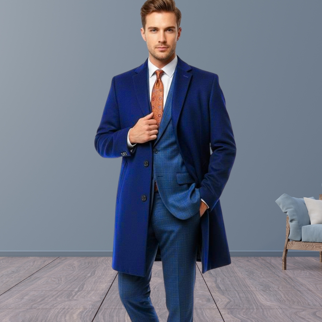 Ultimate Classic Topcoat: Why It Never Goes Out of Style 11 A well-dressed man standing indoors, wearing a blue overcoat over a blue suit with a white shirt and orange patterned tie. He poses confidently with one hand adjusting his coat. The background is a neutral gray, with a wooden floor and a glimpse of a chair with cushions on the right.