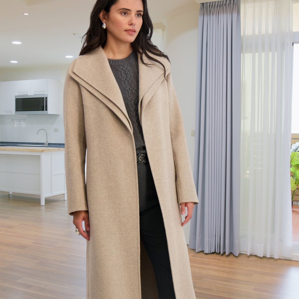 Woman wearing a tan long wool coat over a dark knit sweater and black pants, standing in a bright modern living space with wooden floors and a white kitchen in the background.