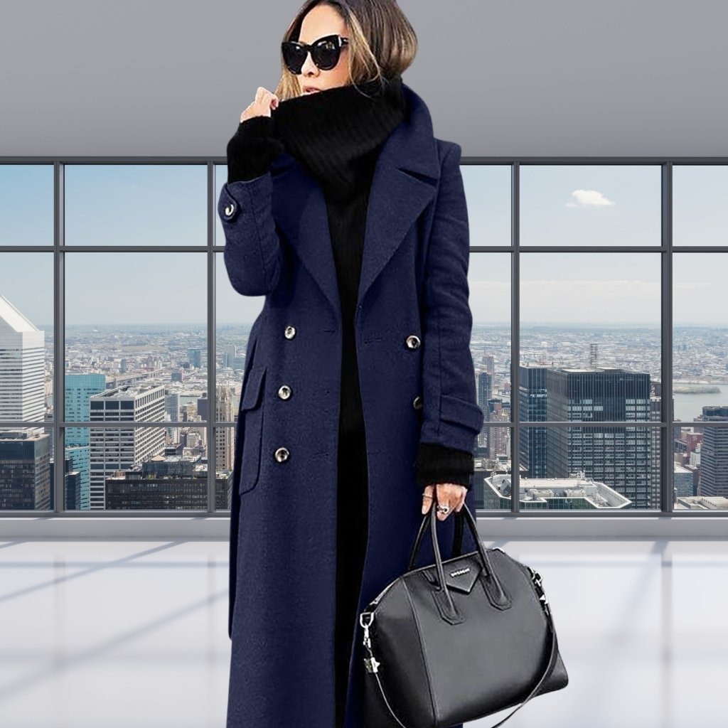 A stylish woman stands in a bright indoor space with a panoramic city view through large windows. She wears a long navy blue double-breasted coat, black turtleneck, black gloves, oversized sunglasses, and carries a black structured handbag.