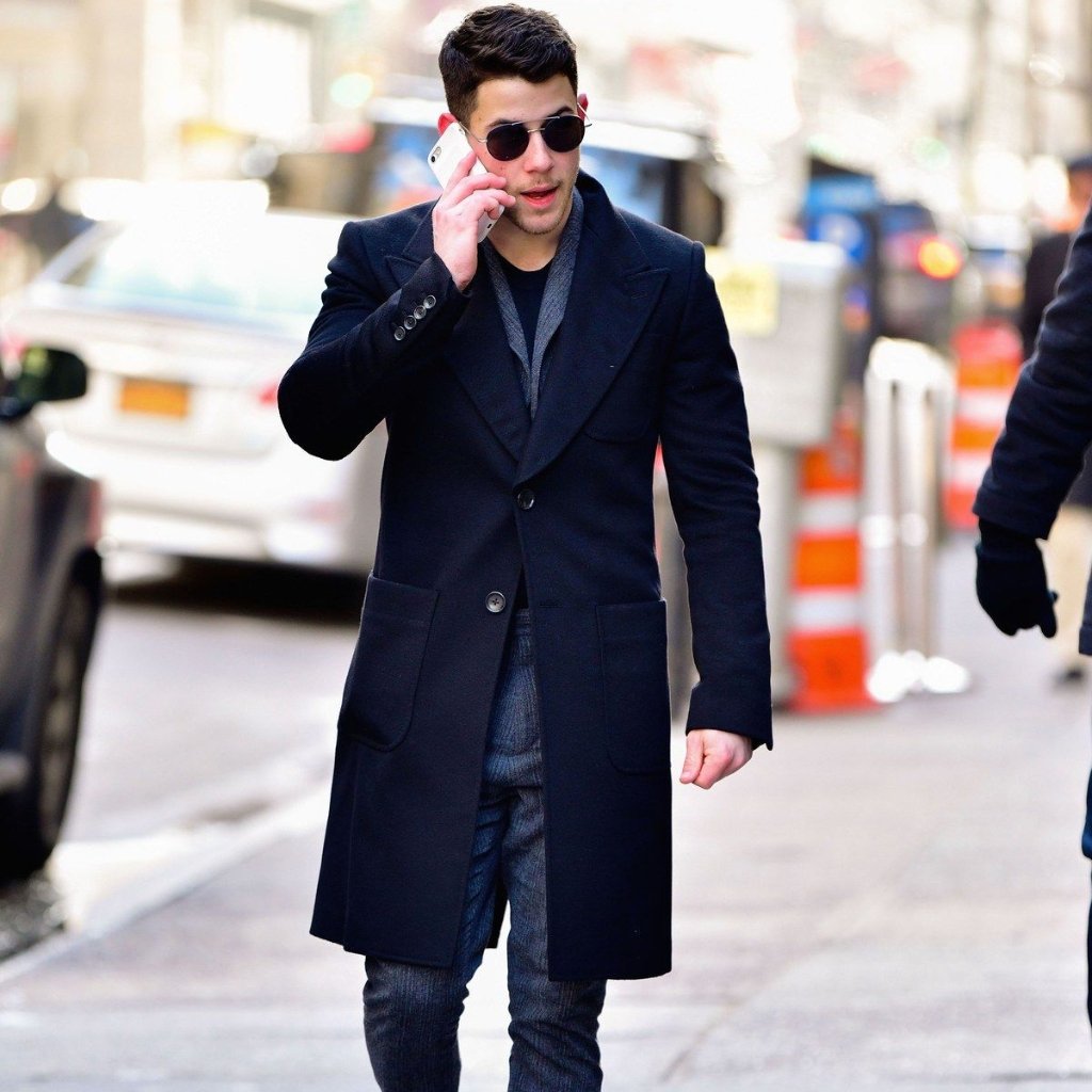 A man walking on a city street wearing a long dark navy coat, sunglasses, and jeans while talking on a smartphone.