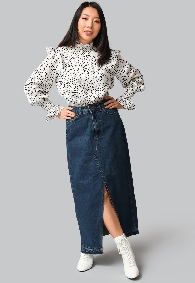 A stylish woman wearing a white long-sleeve blouse with black polka dots and ruffled shoulders, paired with a high-waisted dark denim A-line skirt that has a front slit. She stands with one hand on her hip, wearing white lace-up ankle boots, against a light gray background. Denim A-Line Skirt