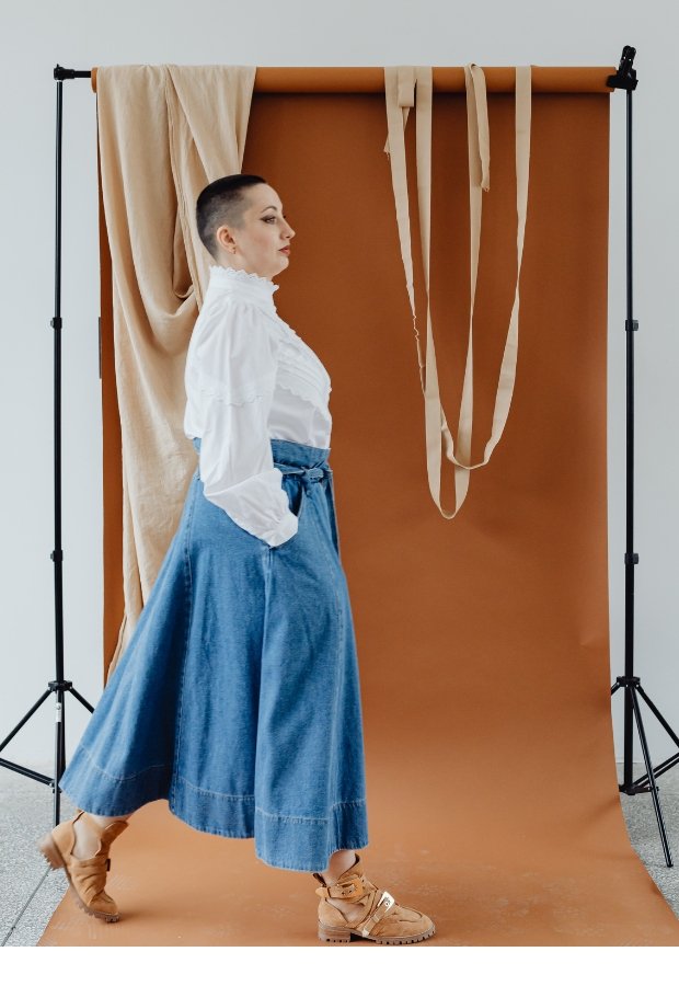 A person with a cropped haircut wearing a white high-neck blouse and a long, blue denim A-line skirt with a sash belt. They are posing in a studio backdrop with a brown seamless paper roll and beige fabric strips hanging in the background, suggesting a fashion shoot emphasizing denim sustainability and timeless silhouettes.