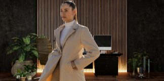 Elegant Topcoat Timeless Statement of Style & Sophistication A stylish woman stands in a modern interior, wearing a light beige overcoat and a white turtleneck. She has her hands in her coat pockets and looks off to the side. The background features a dark wall with wooden slats, a desk with a computer, and several potted plants. Elegant Topcoat