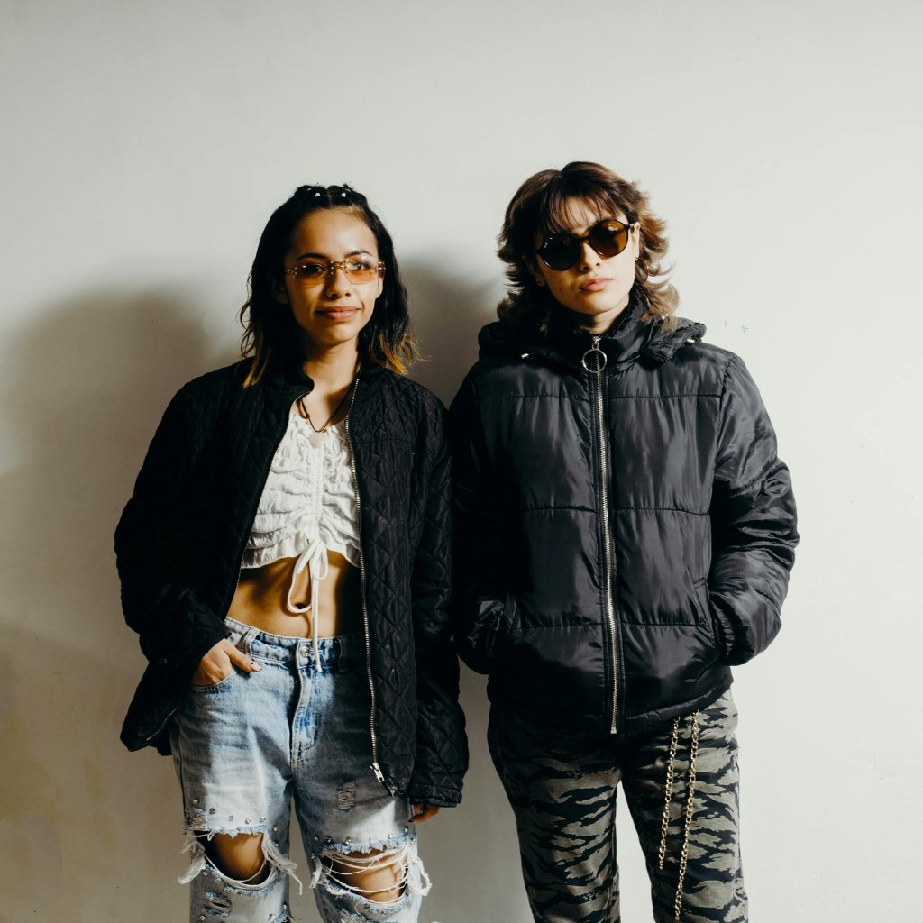 Two stylish young adults stand side by side against a plain light wall, wearing dark puffer jackets and casual distressed outfits. The person on the left wears a black quilted jacket, a white cropped top, and ripped denim shorts; the person on the right wears a black puffer jacket, camouflage pants, and sunglasses.