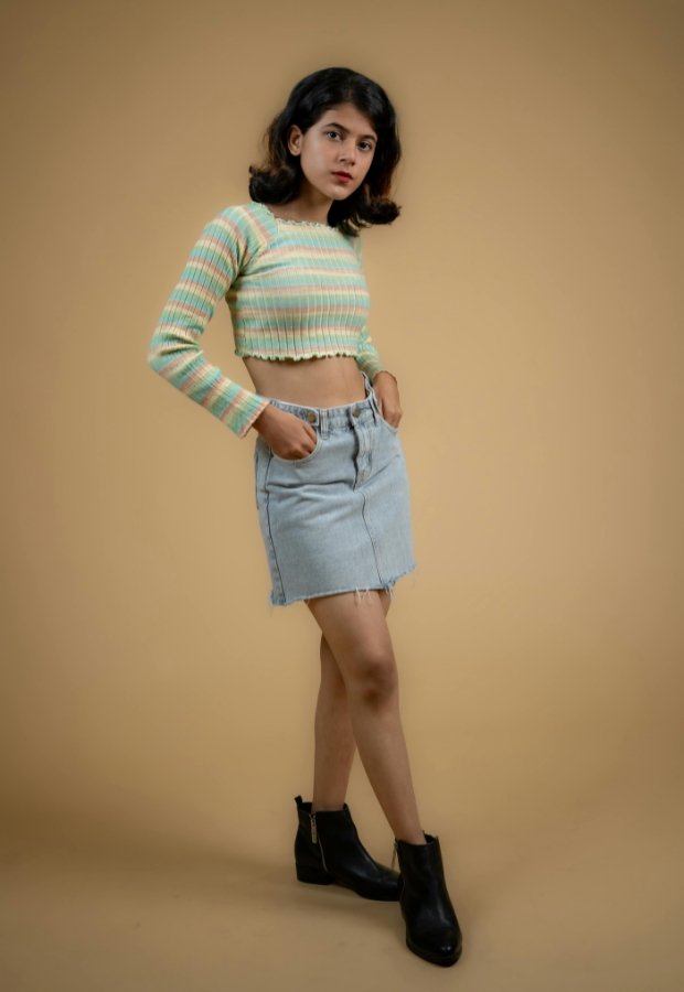 A stylish young woman stands against a warm beige background, wearing a pastel striped cropped knit top, a light-wash denim A-line skirt, and black ankle boots. She has one hand in her pocket, posing confidently.