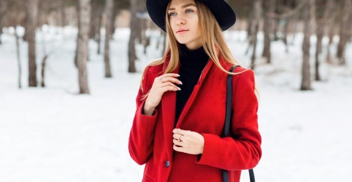 A woman wearing a bright red coat and a black turtleneck stands in a snowy forest, holding a black strap over her shoulder and wearing a black hat. Great Coats for Winter