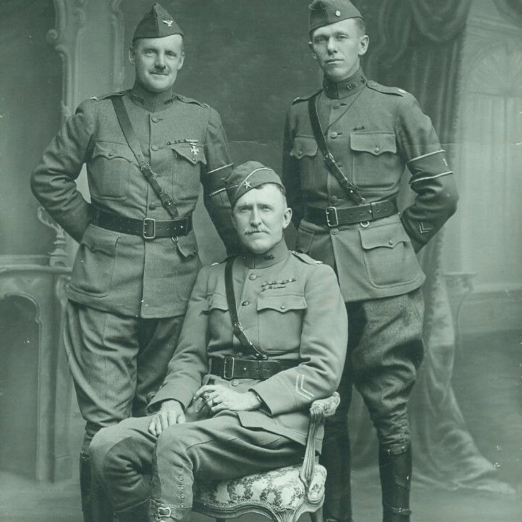 Three World War I-era soldiers in uniform posed for a formal photograph; one seated in a carved chair, two standing on either side.