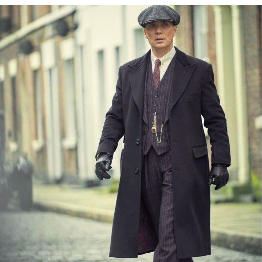 Man walking on a cobblestone street wearing a vintage 1920s-style black overcoat, pinstripe three-piece suit, flat cap, gloves, and a pocket watch chain visible.