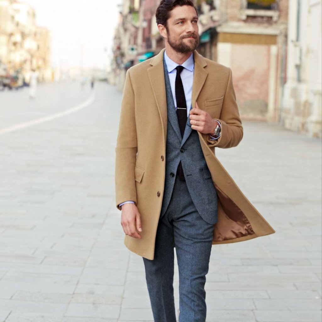 A man walking outdoors wearing a tan overcoat over a gray three-piece suit with a black tie; he looks off to the side and adjusts his coat as it billows slightly behind him.