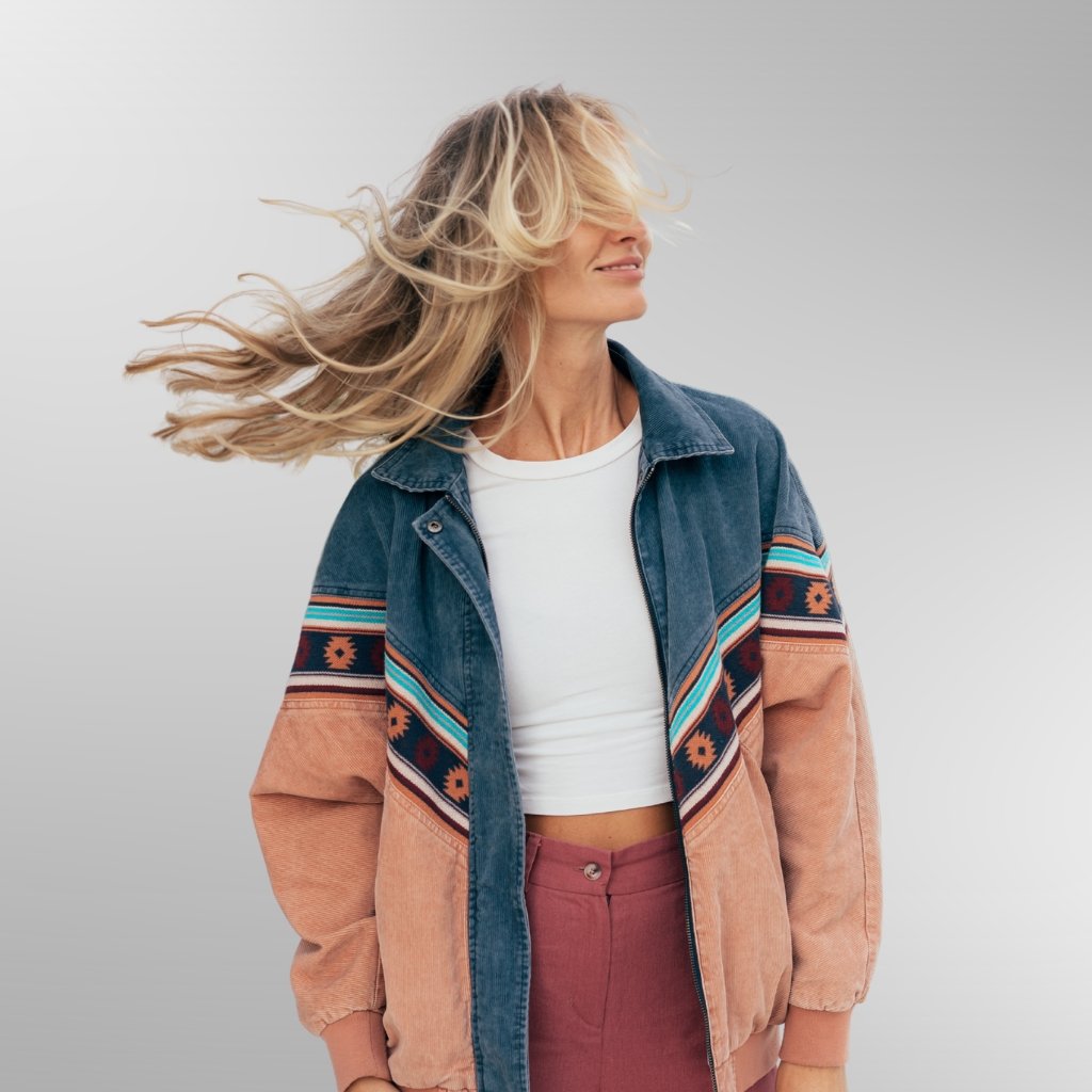 A stylish woman wearing a color-blocked jacket with desert tones and geometric patterns, white crop top, and high-waisted pinkish-red pants. Her blonde hair is tied back and blowing in the wind against a light gray gradient background.