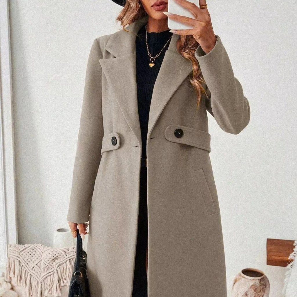 A person wearing a long beige or taupe wool coat with wide lapels and a belt detail, standing indoors. They hold a white phone in front of their face and carry a small black handbag. The outfit is paired with a black top underneath and layered necklaces.