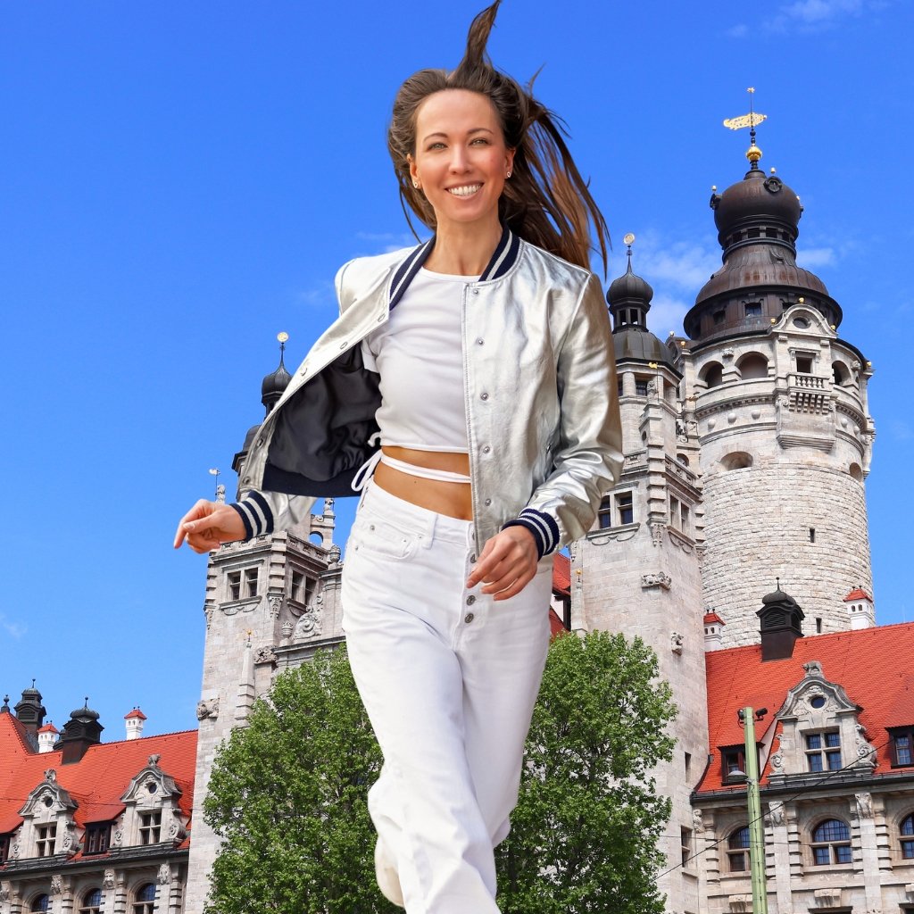 A woman walks in front of a historic castle with tall towers and a blue sky. She wears a silver bomber jacket, white crop top, and white jeans, smiling as she strides forward.