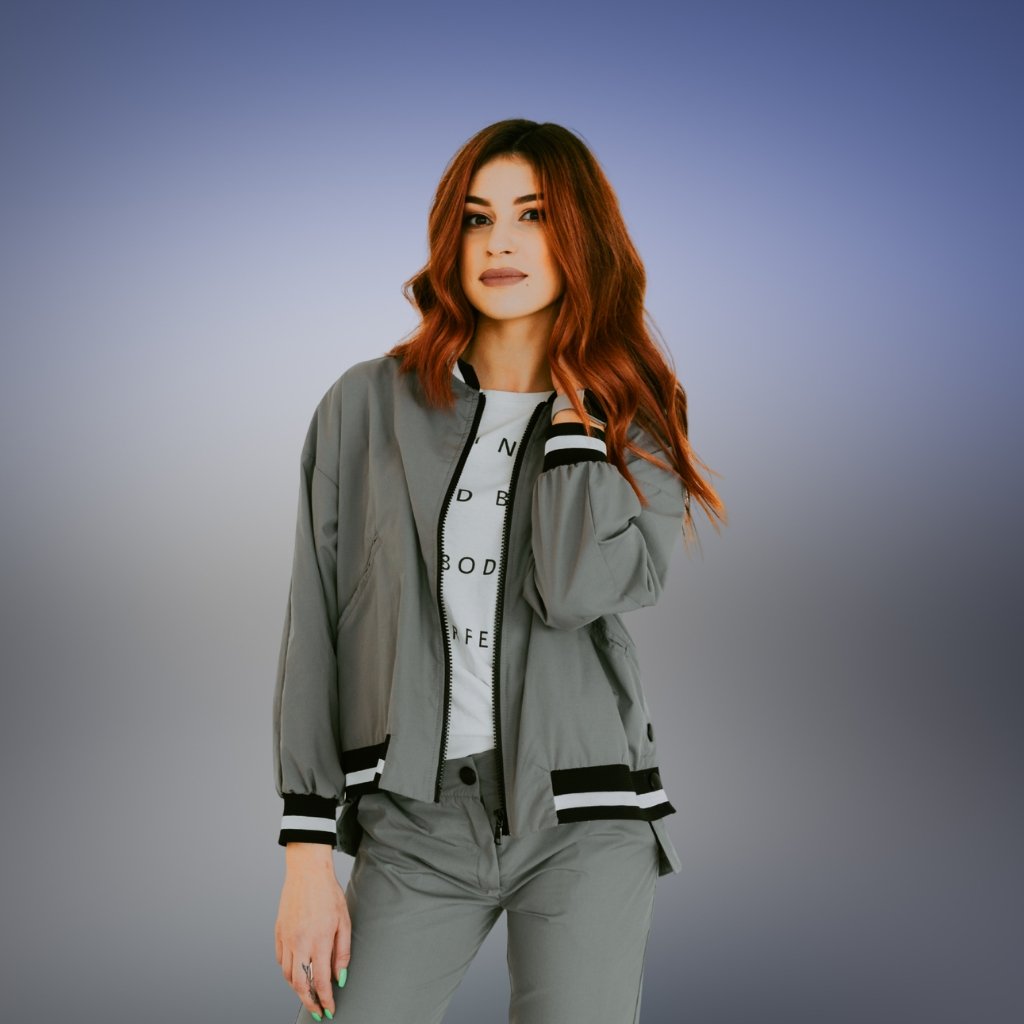A stylish woman with shoulder-length red hair wearing a light gray bomber jacket and matching pants, standing against a soft blue-gray gradient background.