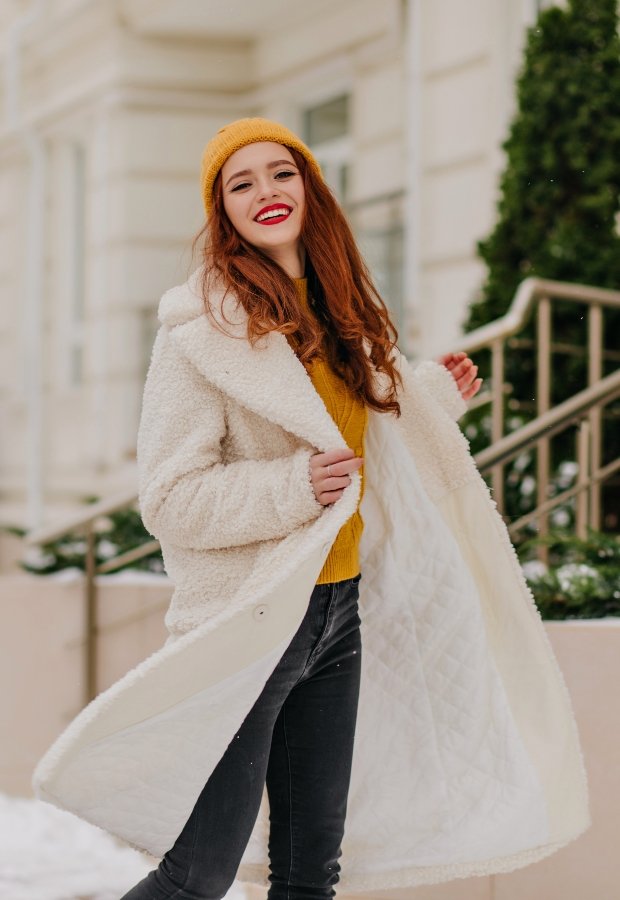 A stylish woman outdoors wearing a long white faux fox fur coat, a mustard knit sweater, black jeans, and a mustard knit beanie, smiling as she walks on a snowy city street.