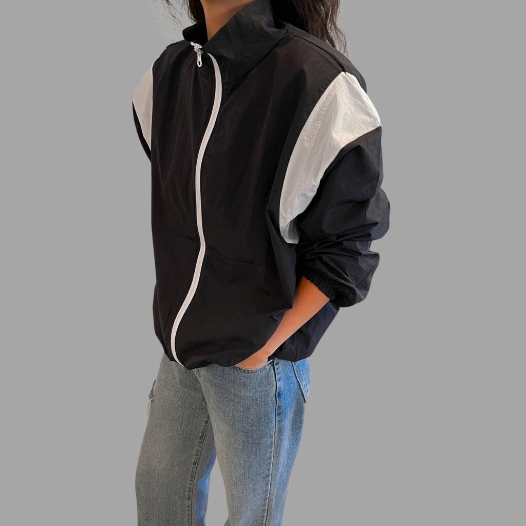 A person wearing a black windbreaker jacket with white accents on the shoulders and a white zipper, paired with light blue jeans. The person’s hands are in their pockets.