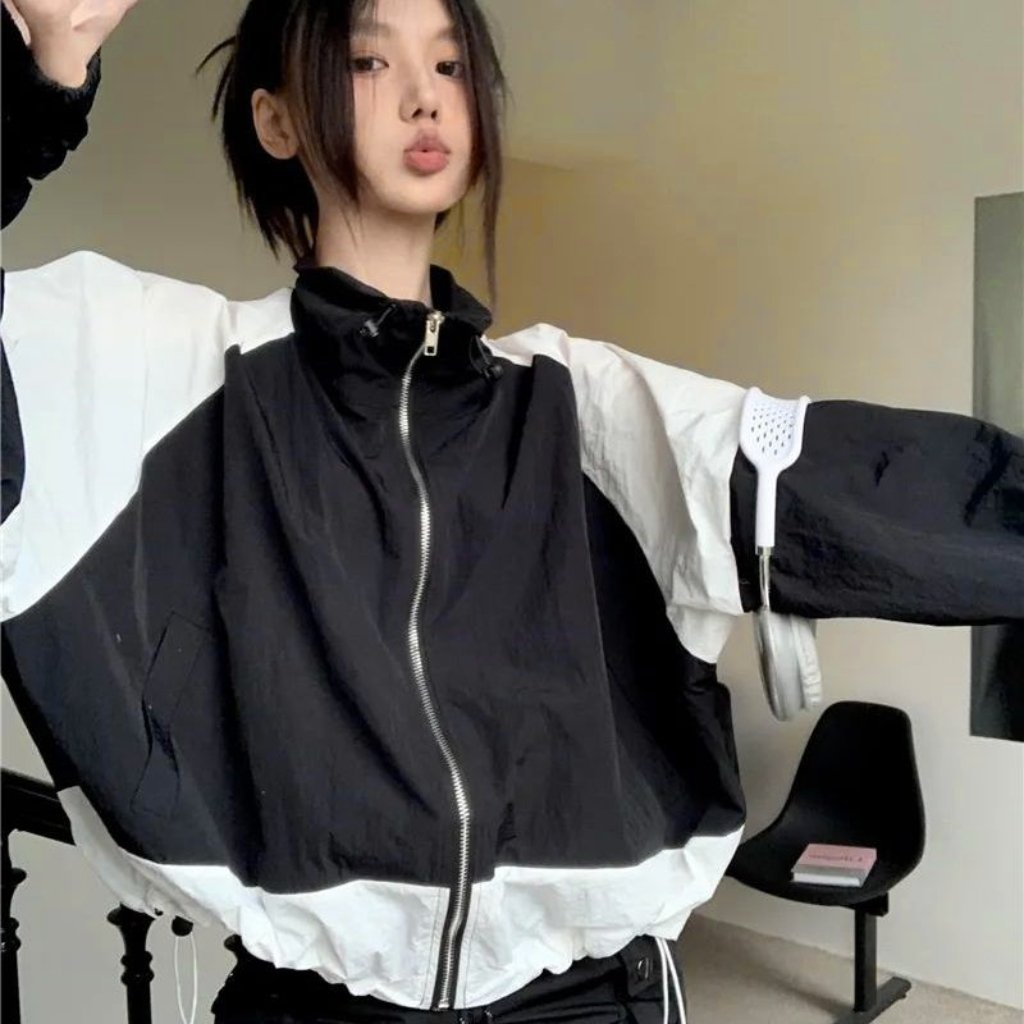 A young woman poses indoors wearing a black and white color-block windbreaker with a zipper front. She extends her arms to the sides, showing the oversized fit and puffed sleeves. Her dark hair is tied back in a loose style, and a chair is visible in the background.