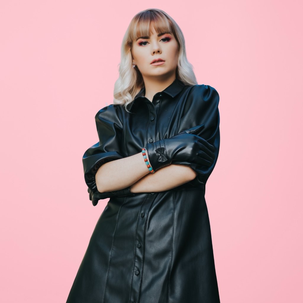 Woman wearing a black longline leather jacket with arms crossed against a solid pink background.
