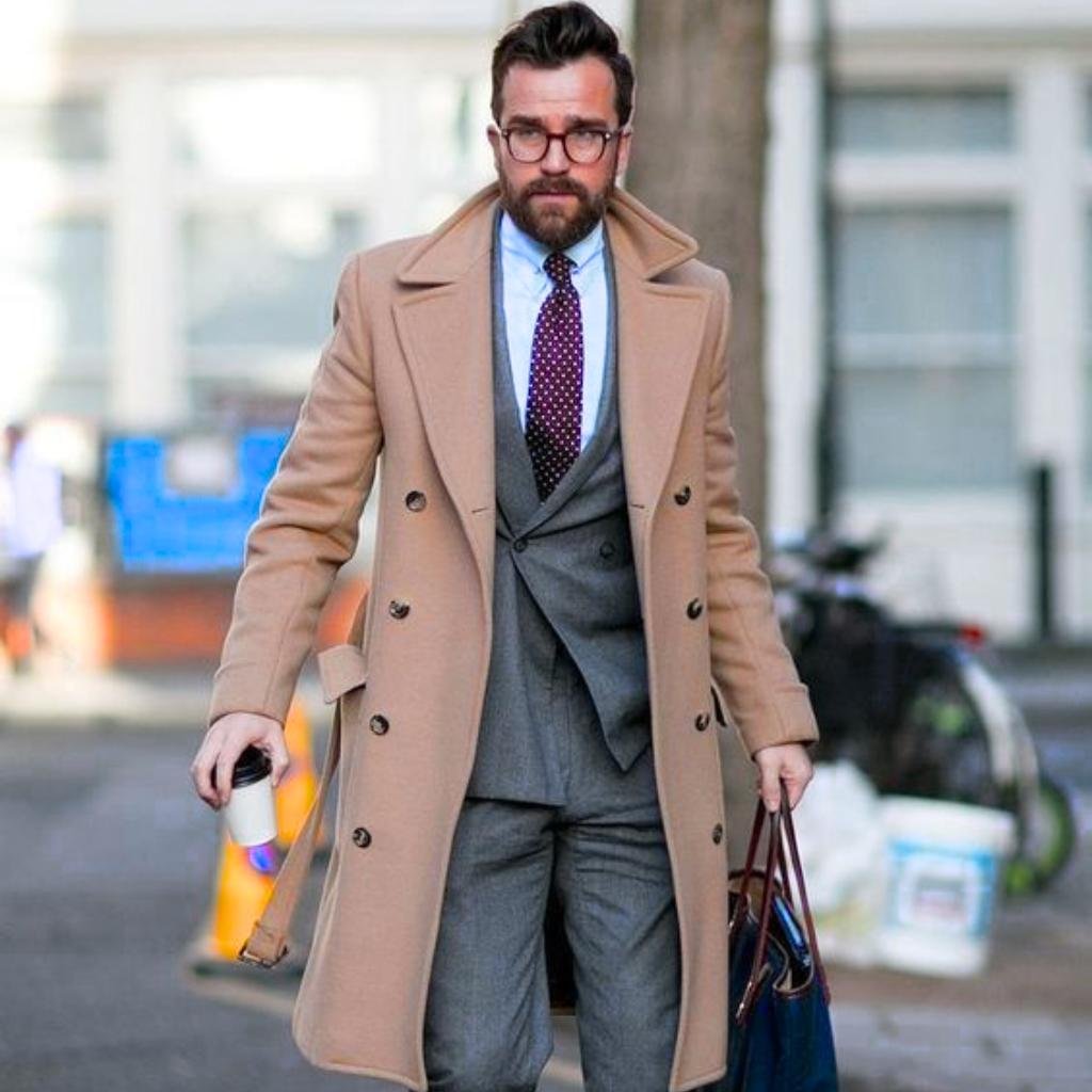 A stylish man with a beard and glasses walks outdoors while holding a coffee cup and a tote bag. He is wearing a tan overcoat over a three-piece gray suit with a blue dress shirt and a purple polka-dot tie.