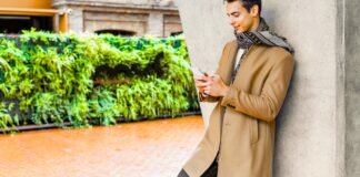 Men’s Topcoat Jackets: Classic Outerwear for Refined Styles. A stylish man leans against a concrete wall outdoors, looking at his phone. He wears a long tan coat and a patterned scarf. Men’s Topcoat Jackets