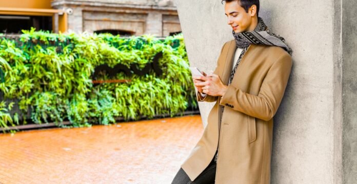 A stylish man leans against a concrete wall outdoors, looking at his phone. He wears a long tan coat and a patterned scarf. Men’s Topcoat Jackets