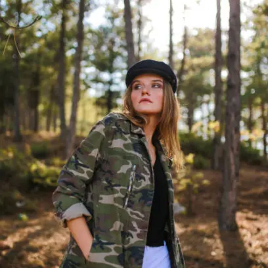 Person wearing a camouflage jacket and black cap, standing in a forest with trees and soft sunlight in the background.Military-Inspired Winter Jackets (11)