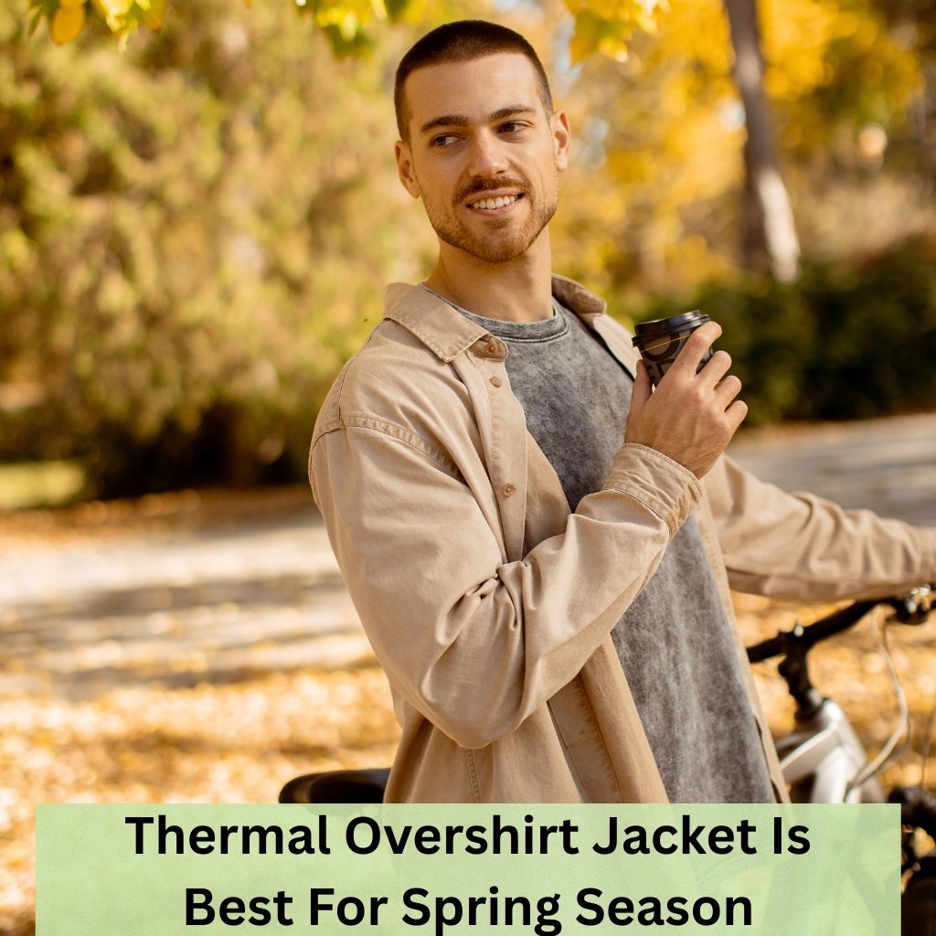 A man outdoors in autumn, wearing a beige overshirt and gray t-shirt, holding a coffee cup near a bicycle; fall leaves in the background.
