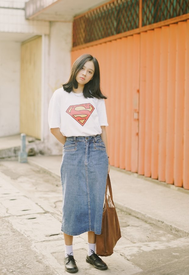 A stylish young woman stands in an urban alley wearing a white t-shirt with a red Superman logo, a mid-length blue denim A-line skirt, black shoes, white socks, and a brown tote bag.