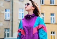 Oversized Track Jacket Women Style Guide Fit Fashion & Trend A woman wearing a colorful retro windbreaker (purple, teal, pink) and sunglasses stands outdoors in front of a beige building. Oversized Track Jacket Women