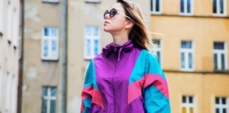 Oversized Track Jacket Women Style Guide Fit Fashion & Trend A woman wearing a colorful retro windbreaker (purple, teal, pink) and sunglasses stands outdoors in front of a beige building. Oversized Track Jacket Women