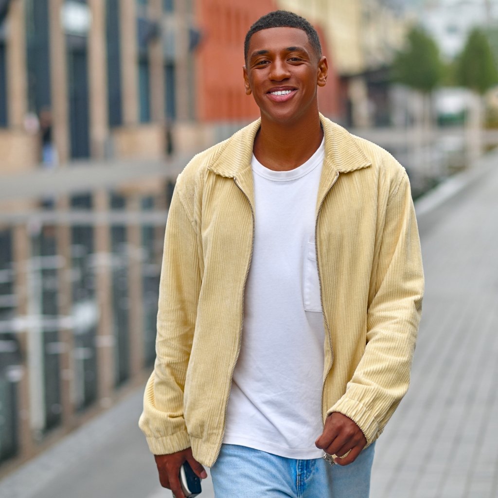 A smiling young man walks outdoors along a city sidewalk, wearing a light beige corduroy jacket, white T-shirt, and light blue jeans.