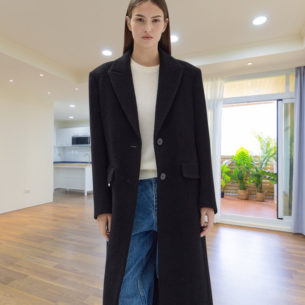 A woman stands indoors wearing a long black coat over a light-colored sweater and blue jeans. The room has wooden floors, bright ceiling lights, and a large glass door leading to a balcony with potted plants in the background.